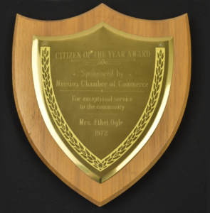 A wooden shield-shaped plaque with a brass plate inscribed "CITIZEN OF THE YEAR AWARD, Sponsored by Mission Chamber of Commerce, For exceptional service to the community, Mrs. Ethel Ogle, 1972," bordered by an engraved leaf pattern.