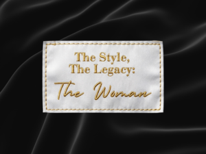 White fabric label with gold embroidered stitching and text reading "The Style, The Legacy: The Woman," displayed against a black satin fabric background.